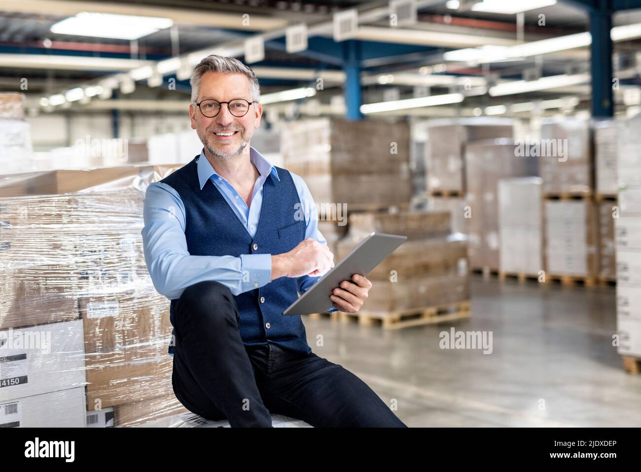 Buon manager che indossa occhiali con tablet PC seduto di fronte alla scatola di cartone in magazzino Foto Stock