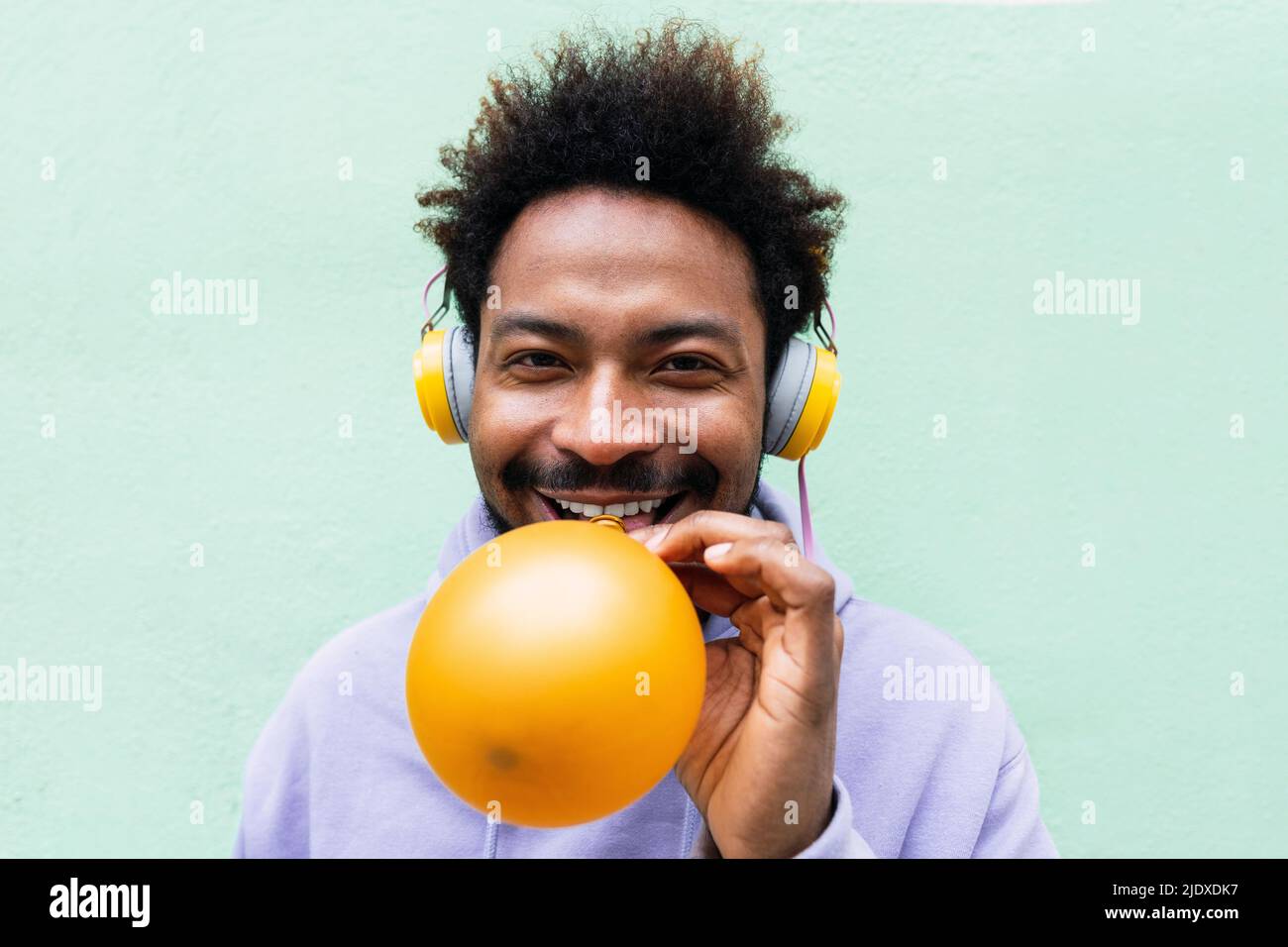 Uomo sorridente che ascolta la musica attraverso le cuffie che soffiano palloncino di fronte alla parete turchese Foto Stock