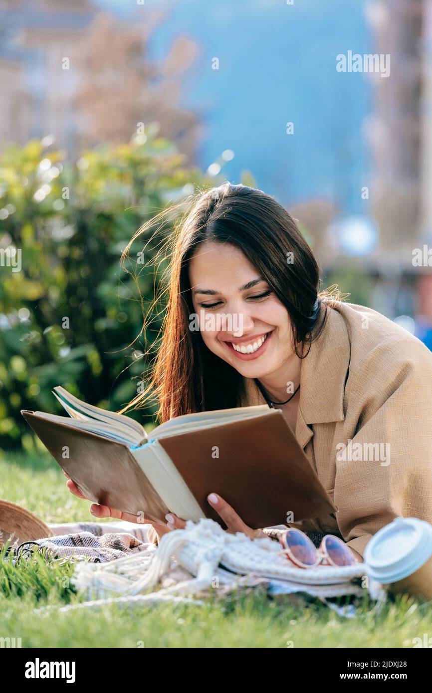 Felice giovane donna con capelli scuri libro lettura sdraiato al parco Foto Stock