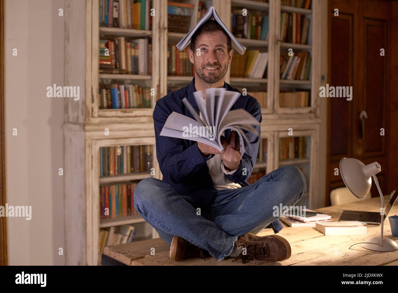 Uomo sorridente che gira pagine di libro seduto sulla scrivania a casa Foto Stock