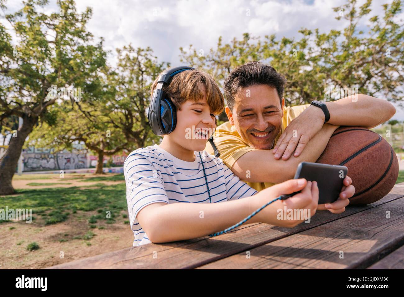 Ragazzo sorridente che ascolta la musica con le cuffie che condividono il telefono cellulare con il padre seduto al tavolo Foto Stock