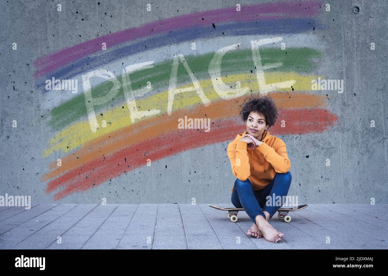 Giovane donna seduta sullo skateboard di fronte alla bandiera arcobaleno con il testo Pace Foto Stock
