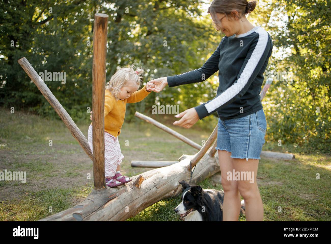 Ragazza che tiene la mano di madre che gioca su albero caduto dal cane nella foresta Foto Stock