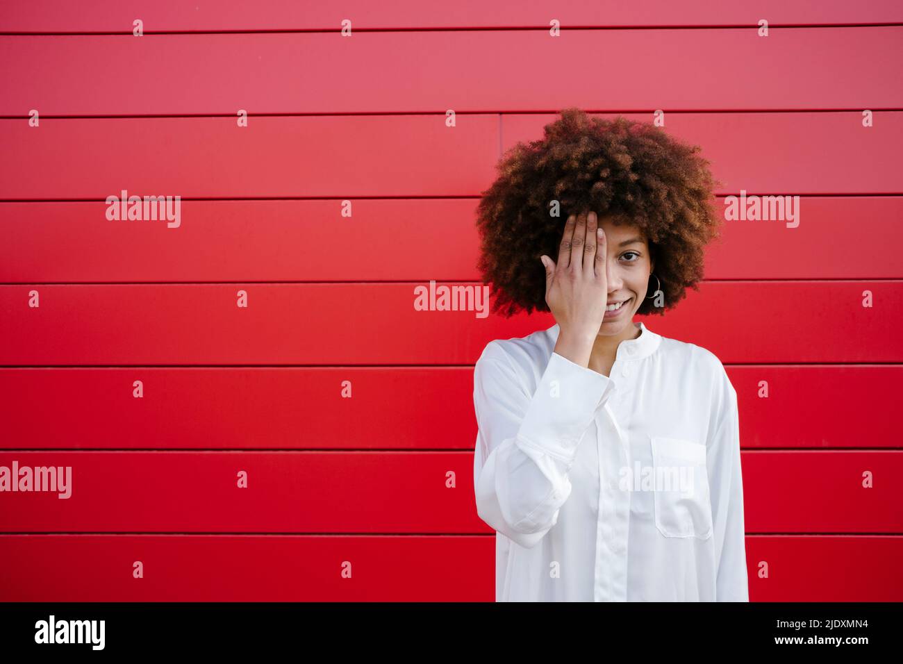 Donna sorridente che ricopre mezza faccia con mano davanti al muro rosso Foto Stock