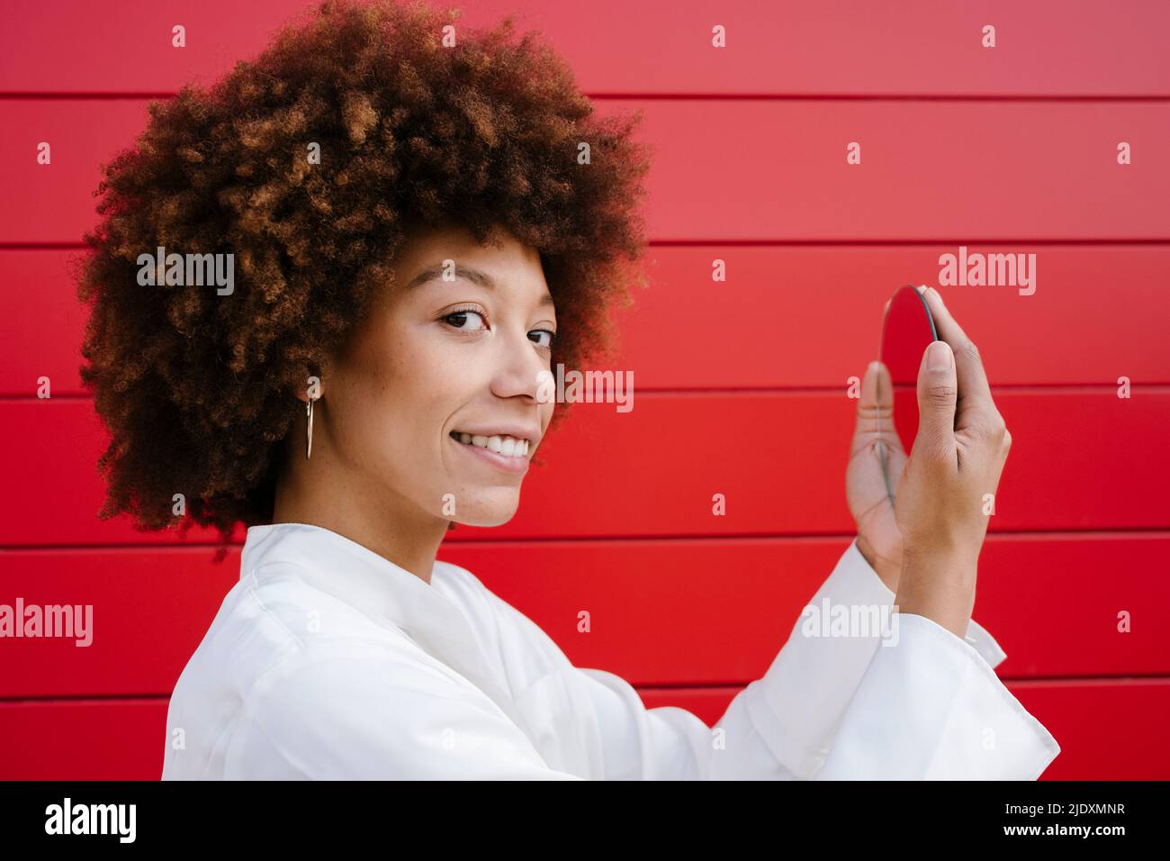 Giovane donna sorridente che tiene lo specchio dal muro rosso Foto Stock