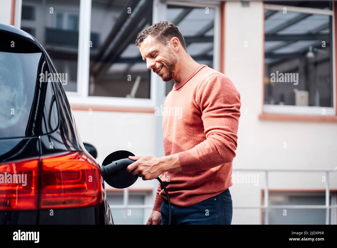 Uomo sorridente che carica l'auto elettrica all'esterno dell'edificio Foto Stock