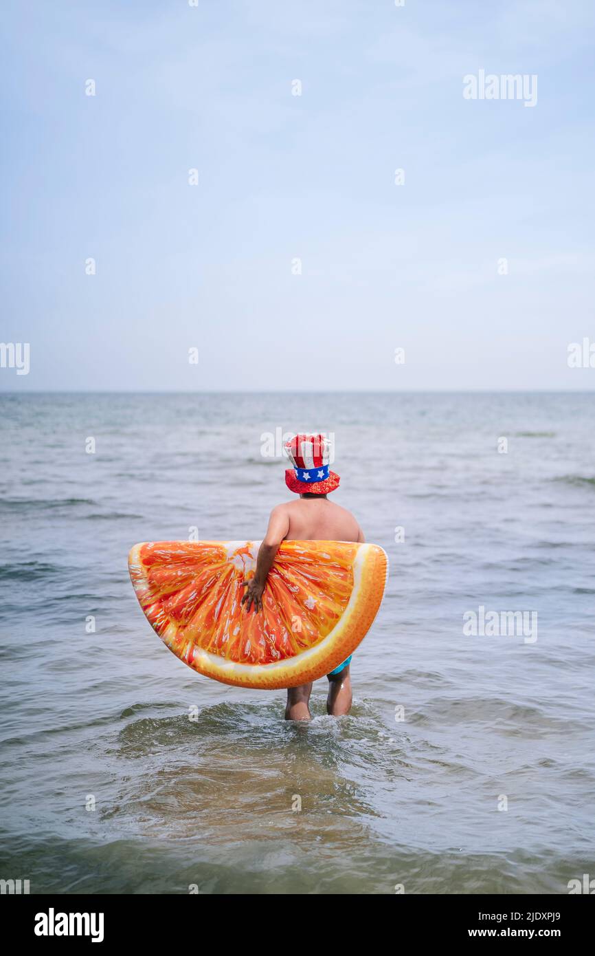 Uomo che porta una fetta gonfiabile di arancia che cammina in mare Foto Stock