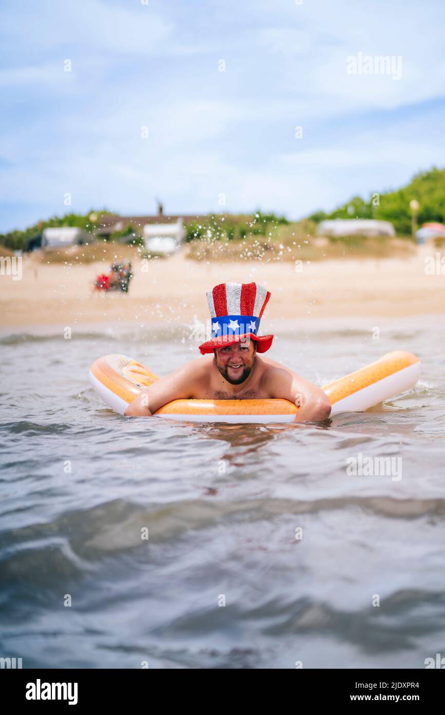 Uomo sorridente che indossa lo zio Sam cappello che nuota con zattera da piscina in mare Foto Stock