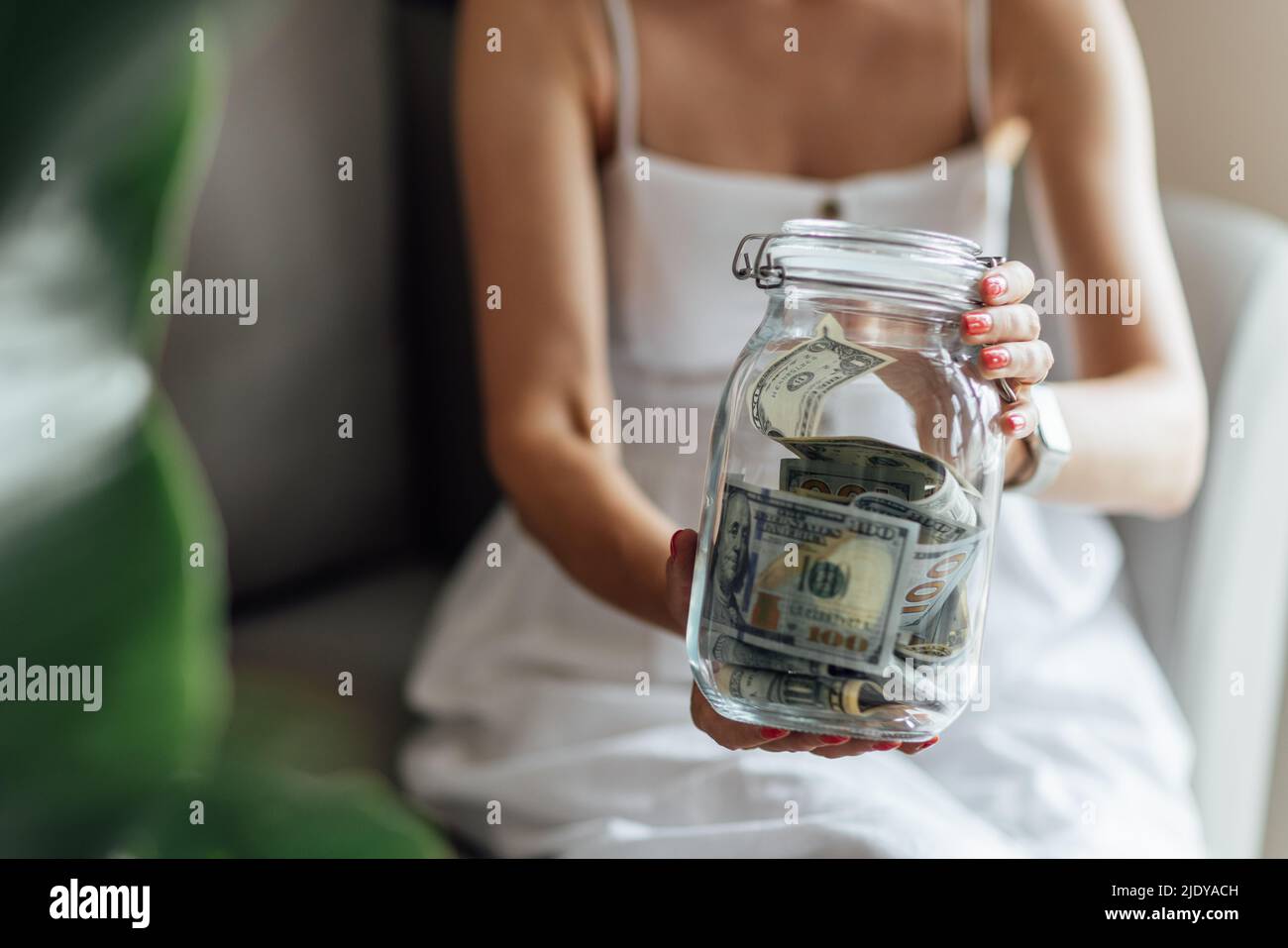 Giovane donna caucasica che tiene una grande bottiglia di vetro con denaro contante bolletta in dollari su un tavolo di marmo bianco a casa. Investimento, previdenza, finanza e. Foto Stock