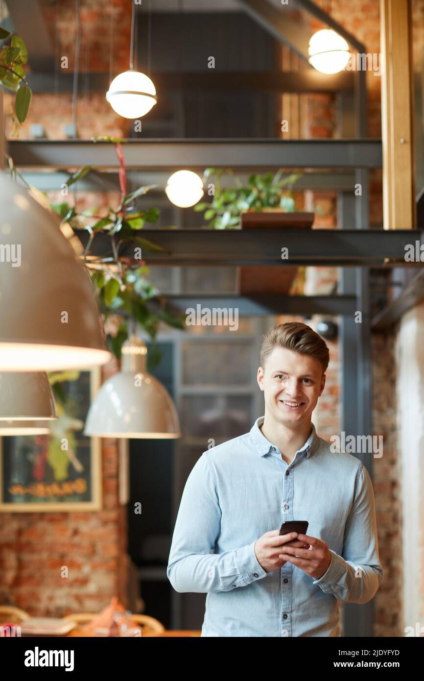 Ritratto di buon bel giovane tester in camicia blu chiaro in piedi nel bar loft e utilizzando smartphone Foto Stock