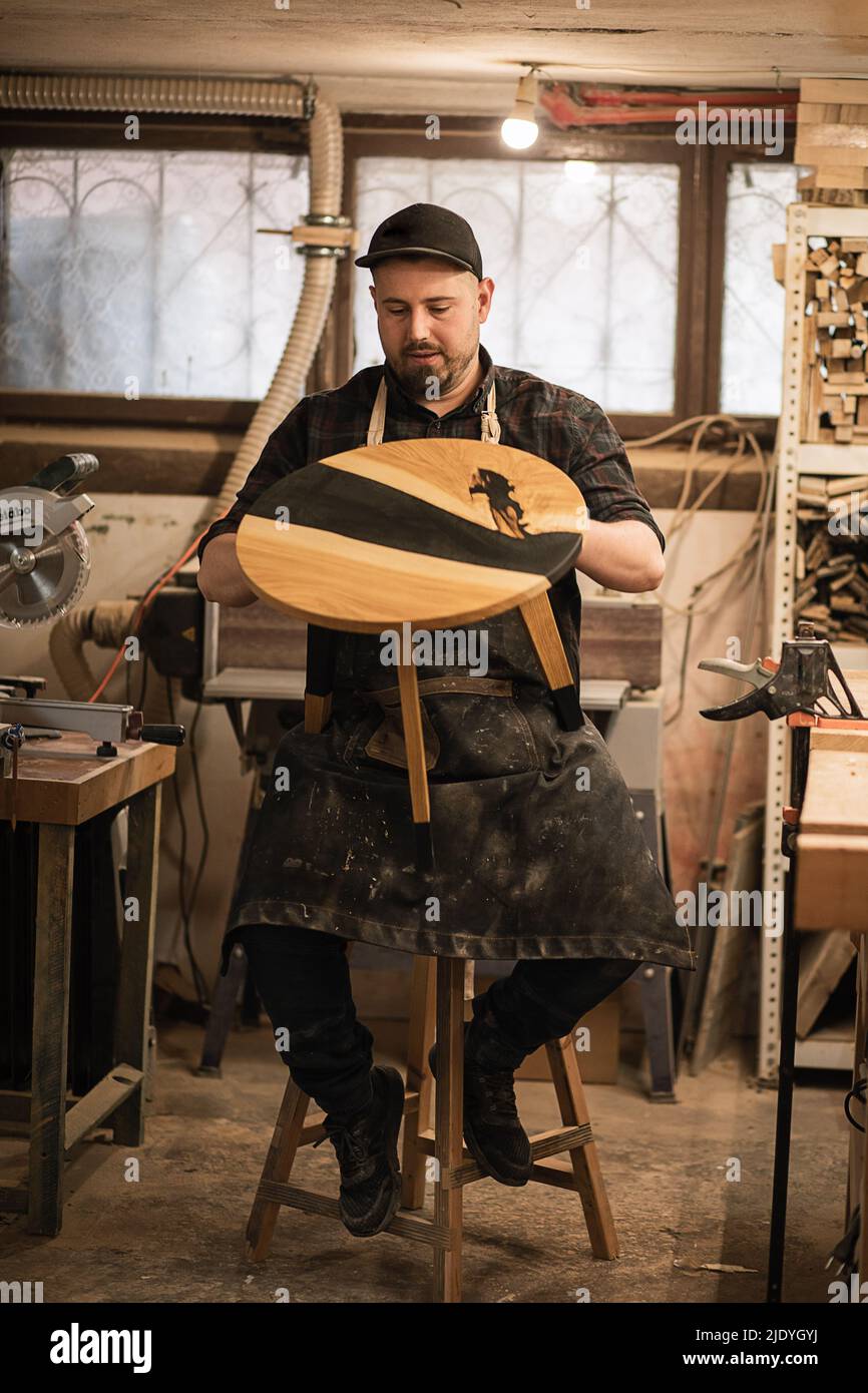 Falegname verticale sicuro e concentrato, carpentiere in uniforme sporca e tappo in garage, officina, controllo tavola rotonda Foto Stock