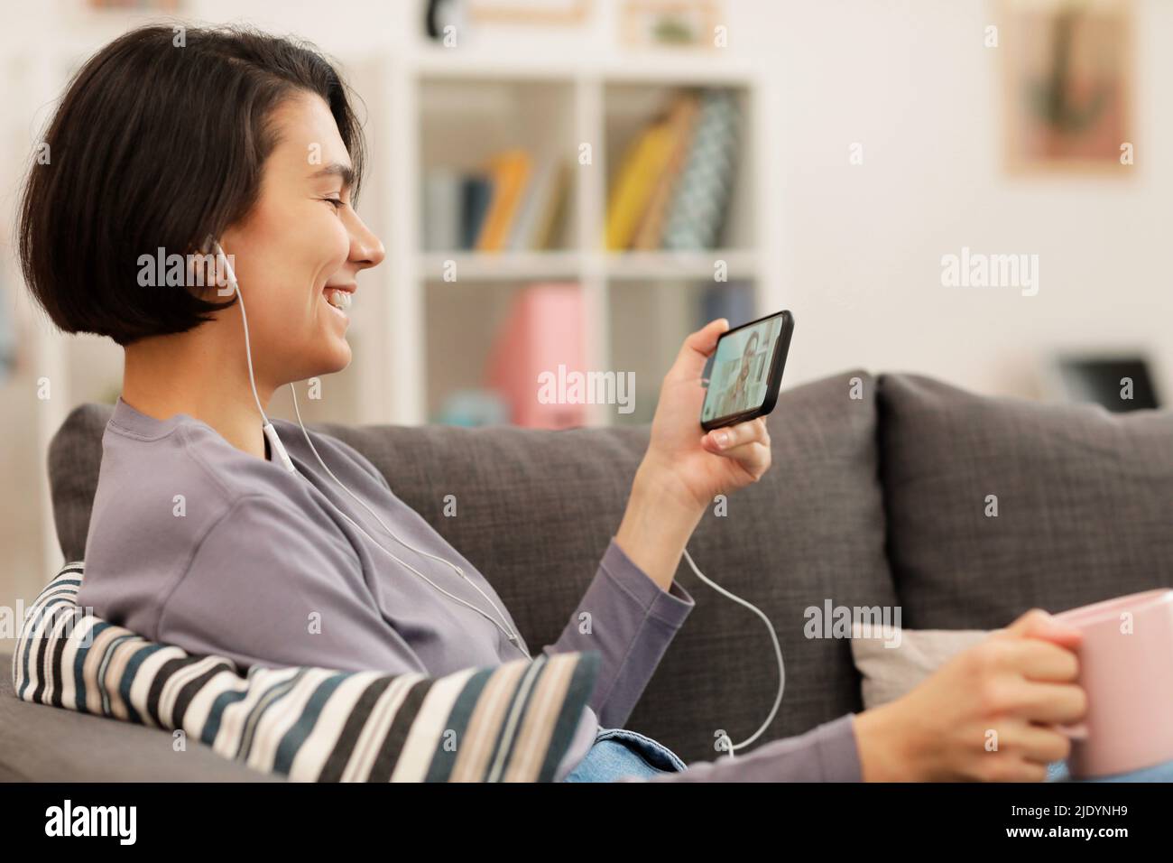 Bella donna giovane positiva con capelli corti seduti sul divano a casa e utilizzando l'app per videoconferenze per la comunicazione Foto Stock