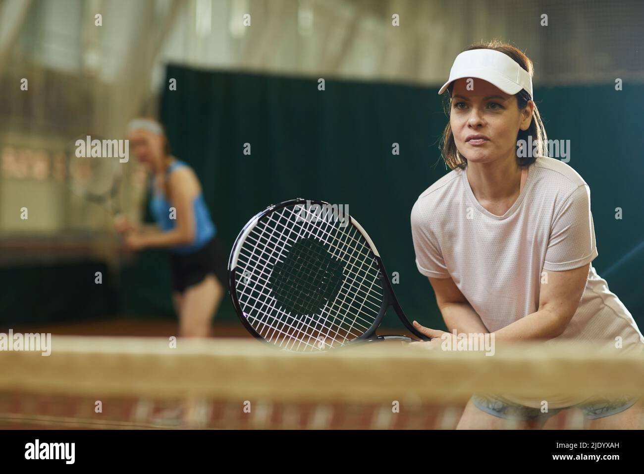 Contenuto entusiasta giovane donna in cappellino da tennis accovacciato in posizione pronta e racket in attesa di servire palla da concorrente Foto Stock