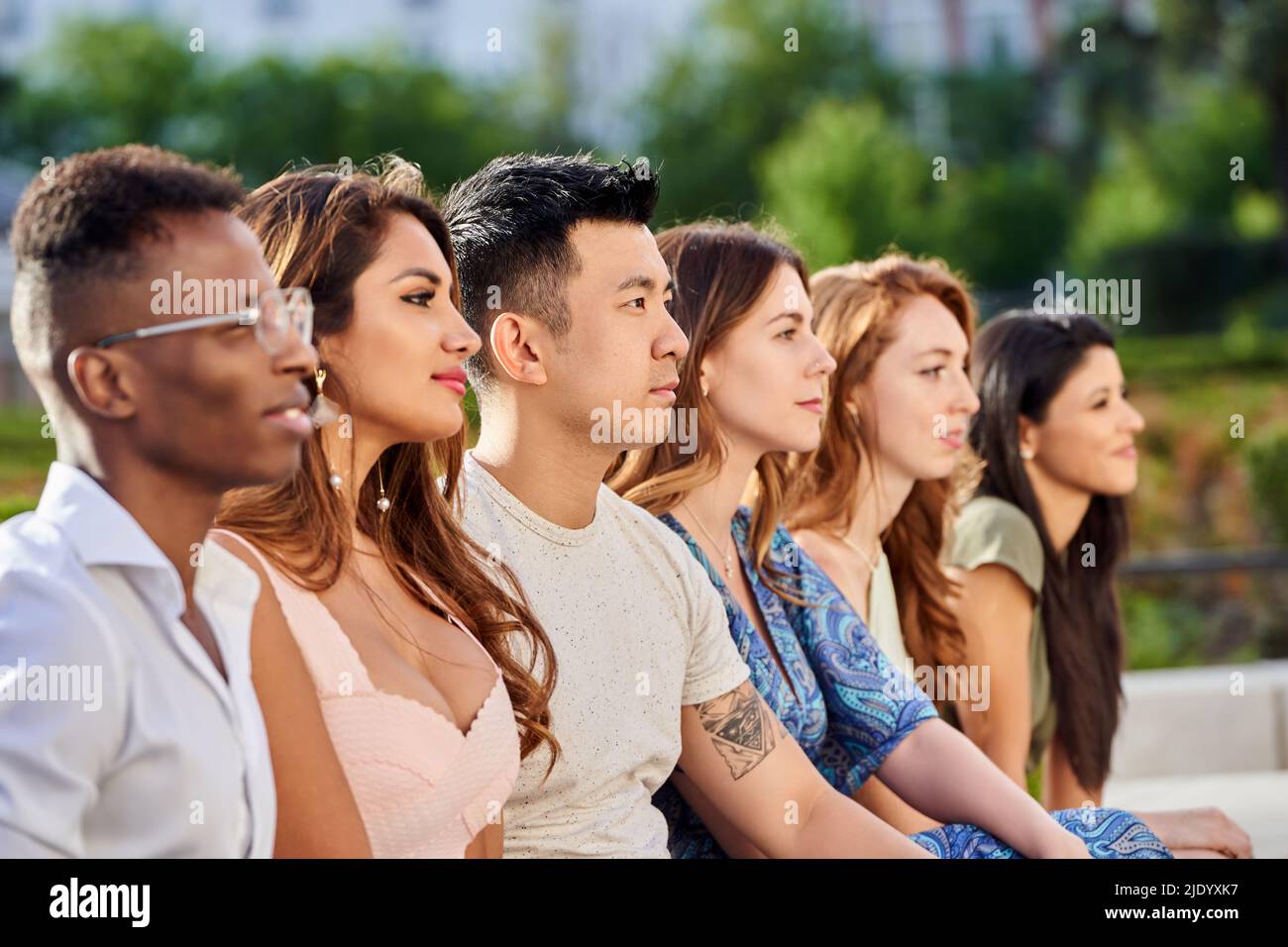 Gruppo di giovani multirazziali che guardano via mentre si siedono in fila insieme all'aperto. Concetto di squadra e unità. Foto Stock