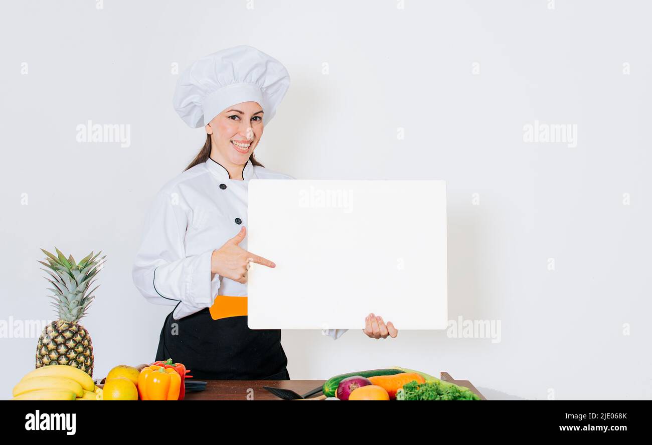 Bella donna chef con tavolo di verdure che tiene un bordo vuoto, Chef donna con verdure al tavolo che tiene menu vuoto. Ragazza chef in cucina Foto Stock