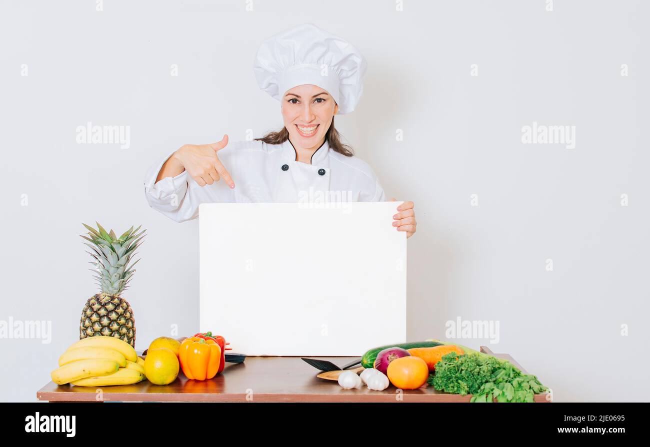 Chef ragazza con verdure al tavolo che tiene un menu vuoto. Bella donna chef con tavolo di verdure che tiene un bordo vuoto, ragazza chef in cucina Foto Stock