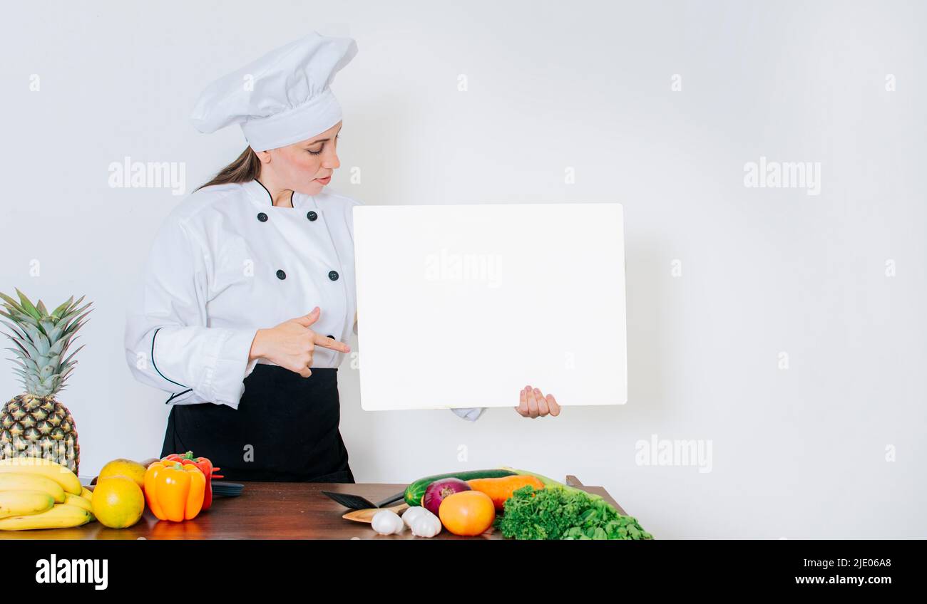 Chef donna con verdure a tavola con menu vuoto. Ragazza chef in cucina mostrando un bordo vuoto, bella donna chef con tavola di verdure Foto Stock