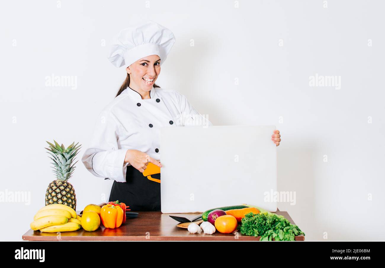 Chef donna con verdure a tavola con menu vuoto. Ragazza chef in cucina mostrando un bordo vuoto, bella donna chef con tavola di verdure Foto Stock