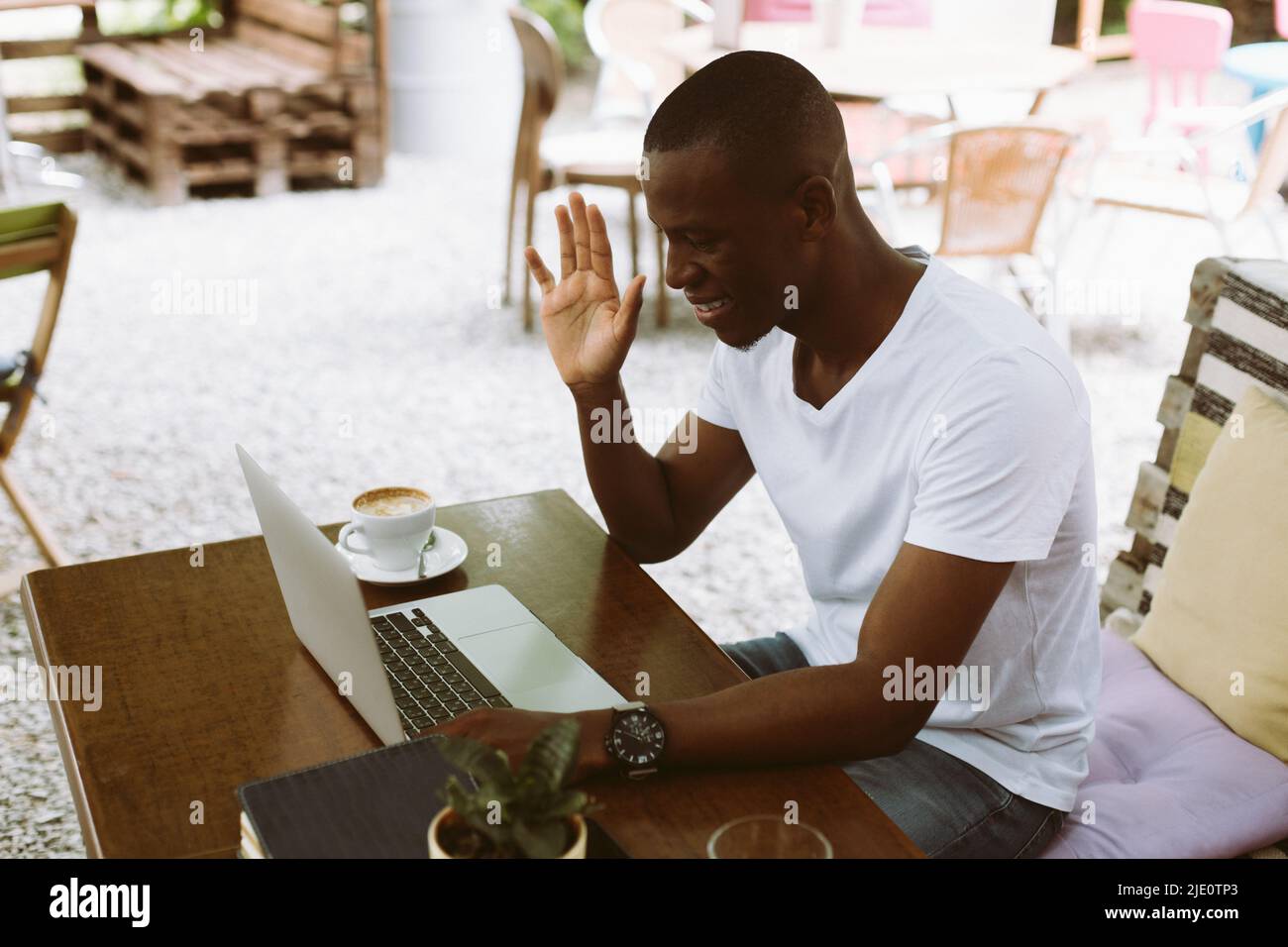 Uomo d'affari multirazziale sorridente e rilassato con notebook, alzando la mano, il saluto, guardare monitor. Ufficio freelance online Foto Stock