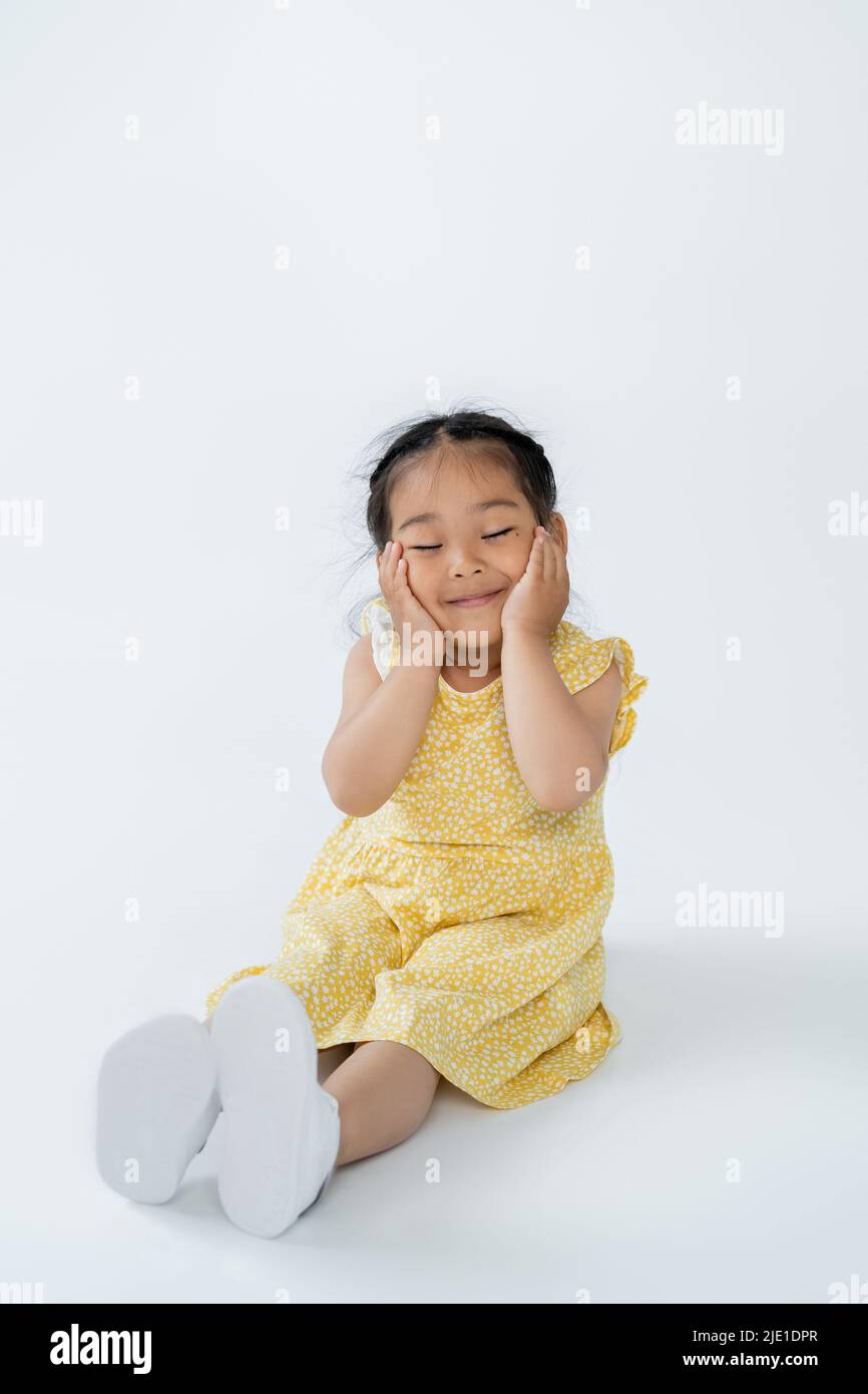intera lunghezza di bambino asiatico carino in abito giallo sorridente con occhi chiusi mentre si siede in grigio Foto Stock