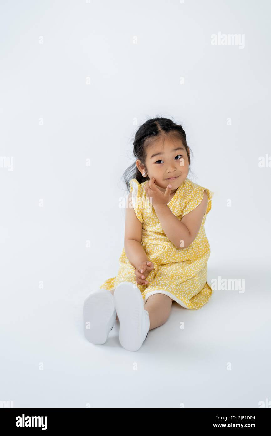 intera lunghezza di bambino asiatico carino in abito giallo seduta su grigio Foto Stock