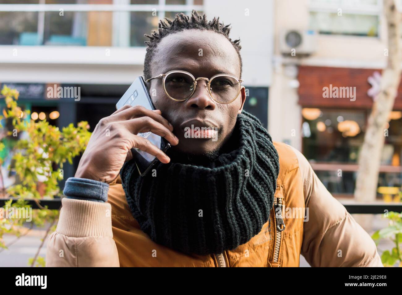 ritratto giovane uomo africano seduto all'aperto con sciarpa nera, giacca color senape e occhiali che parlano al telefono all'aperto con locale commerciale Foto Stock