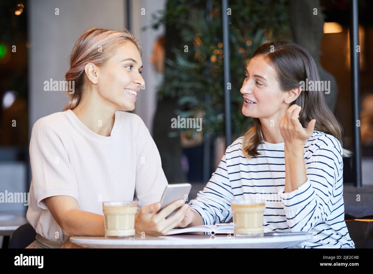 Ragazze attraenti positive in abbigliamento casual seduti a tavola e discutere di informazioni sui social media durante la riunione in caffetteria di notte Foto Stock