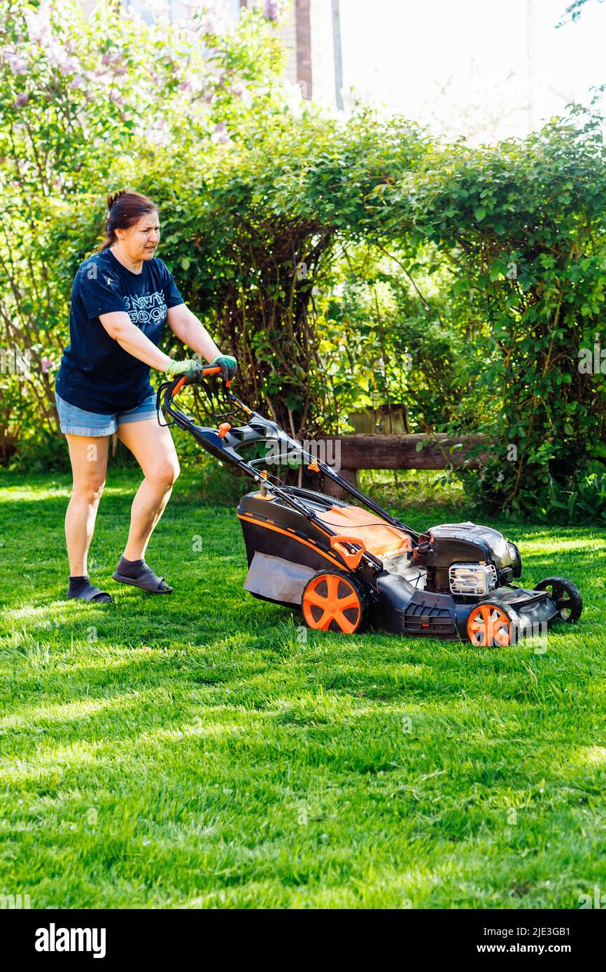 Vista posteriore del giardiniere femminile in abiti casual godendo di taglio erba fresca con rasaerba o taglierina erba nel cortile Foto Stock