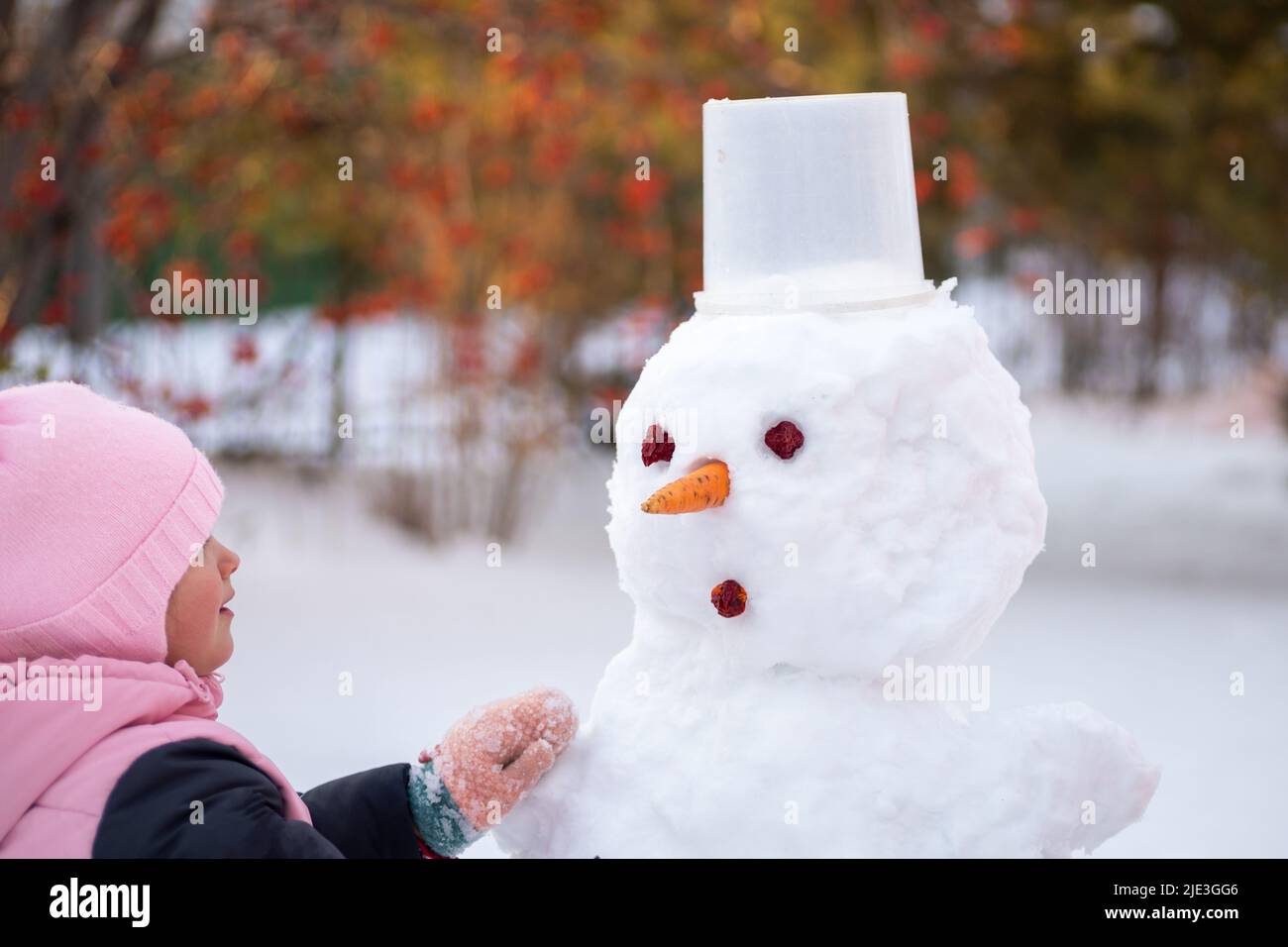 Bambina che fa il viso di un pupazzo di neve con mezzi improvvisati di giorno, mentre si cammina nel parco con alberi e luce del sole sullo sfondo. Genitori Foto Stock