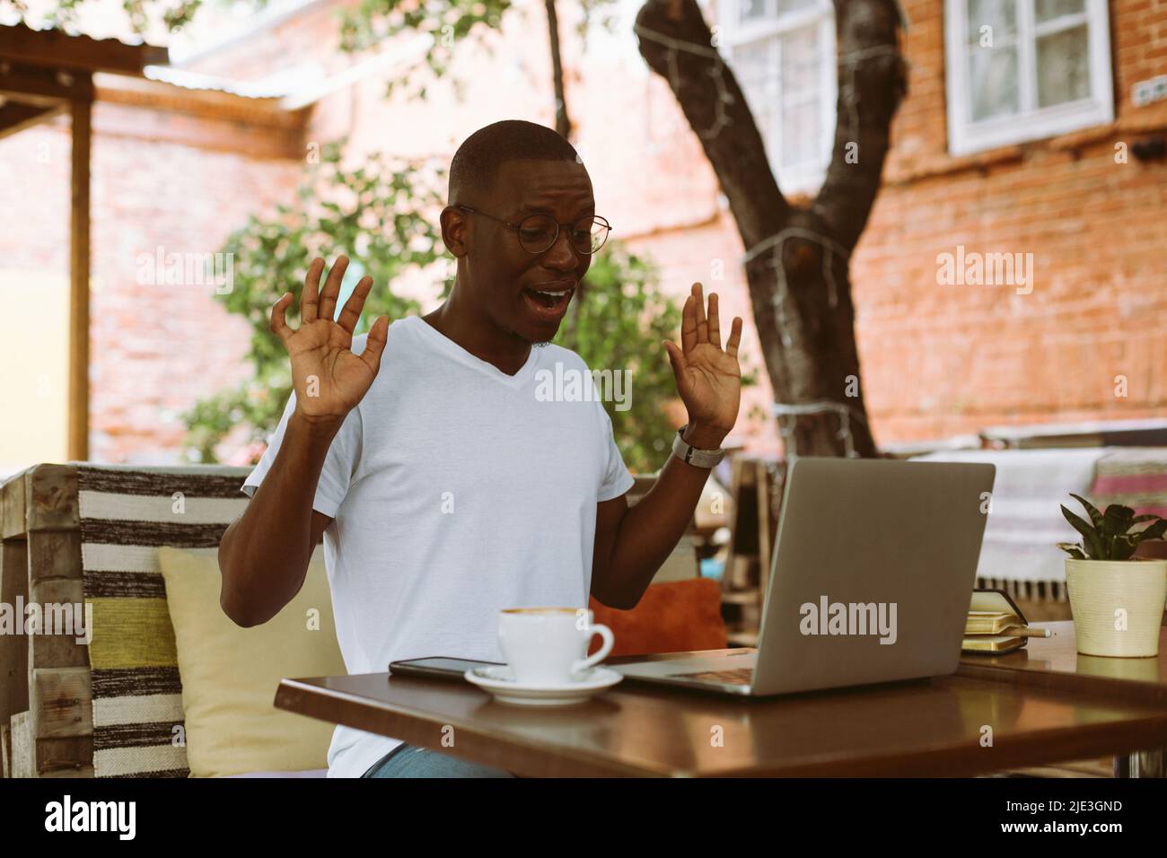 Sorprende che l'uomo afroamericano lavori su un portatile con Internet senza fili, sollevando le braccia in caffetteria. Webinar e-learning Foto Stock