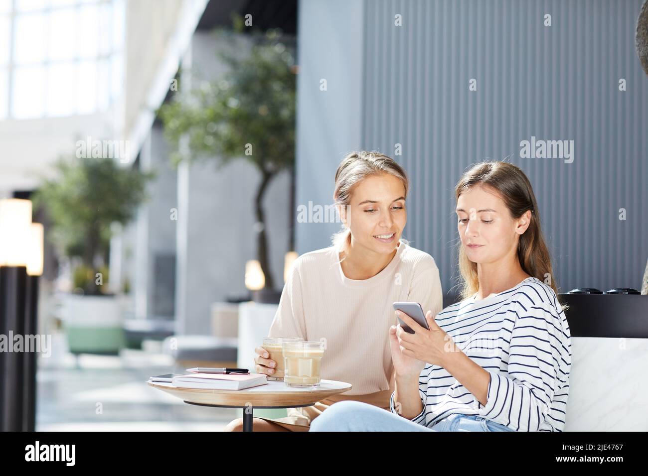 Positive ragazze moderne in abbigliamento casual seduto sul divano e bere caffè mentre surf sociale media insieme Foto Stock