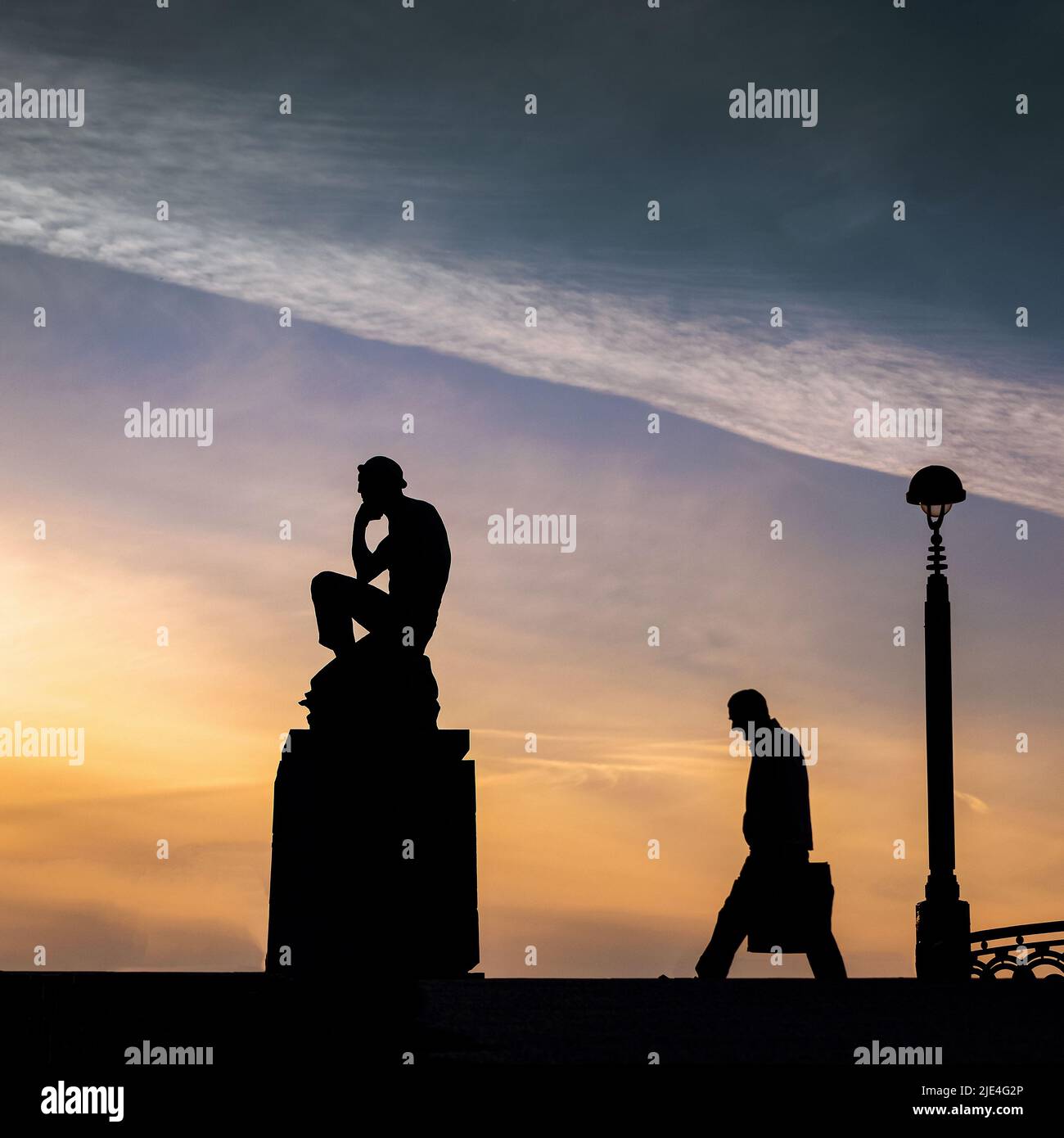 Un uomo d'affari cammina e pensa al tramonto accanto ad una scultura meditativa del vecchio uomo Foto Stock