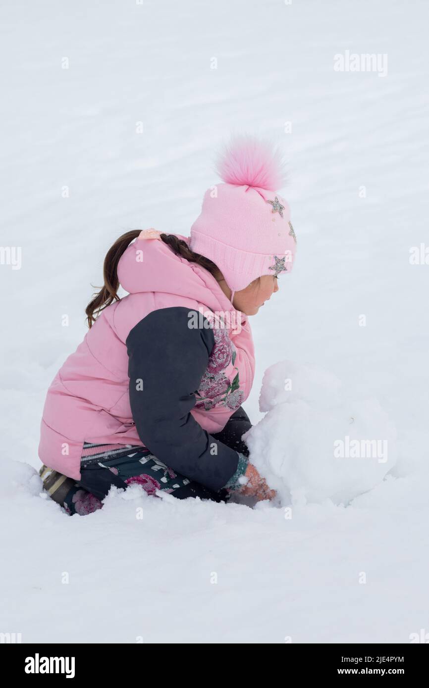 Bambina che fa grande palla di neve per costruire uomo di neve in parco di giorno con terreno coperto di neve in background. I genitori che trascorrono il tempo con Foto Stock