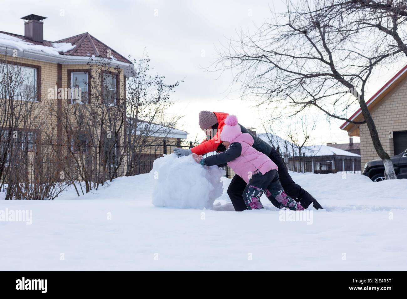 Bambina con madre che fa la palla di neve voluminosa per costruire l'uomo di neve sul cortile in serata con gli alberi e la casa in background. I genitori trascorrono del tempo Foto Stock