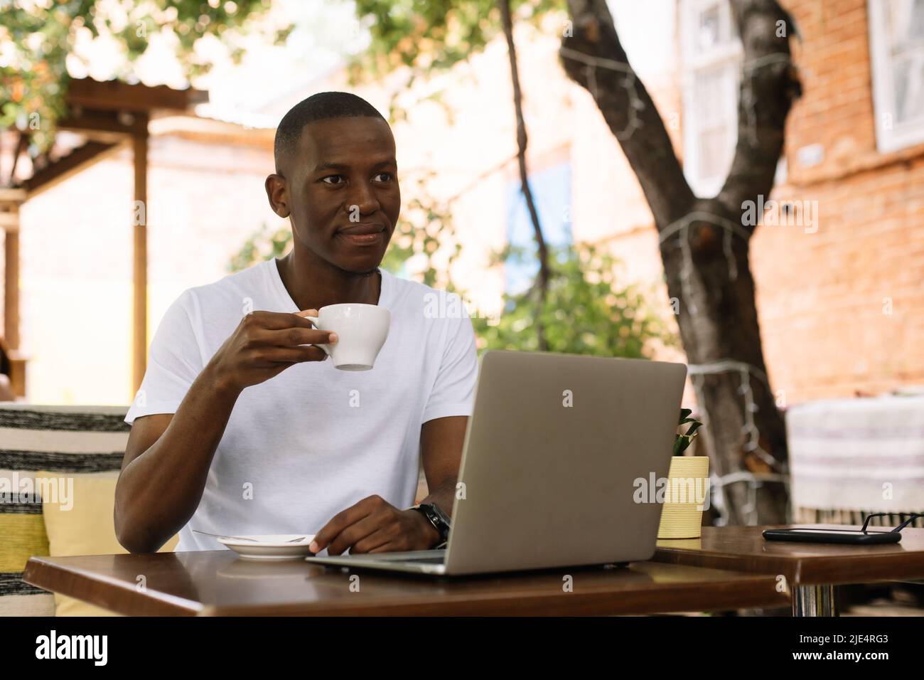 Uomo d'affari multirazziale sicuro e sorridente che beve caffè, lavorando online utilizzando il laptop in un bar all'aperto. Processo remoto Foto Stock