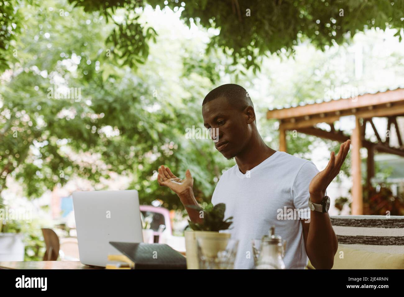 Sorpreso e confuso afroamericano uomo con computer portatile in caffè non sa cosa fare, alzando le mani in un caffè all'aperto Foto Stock