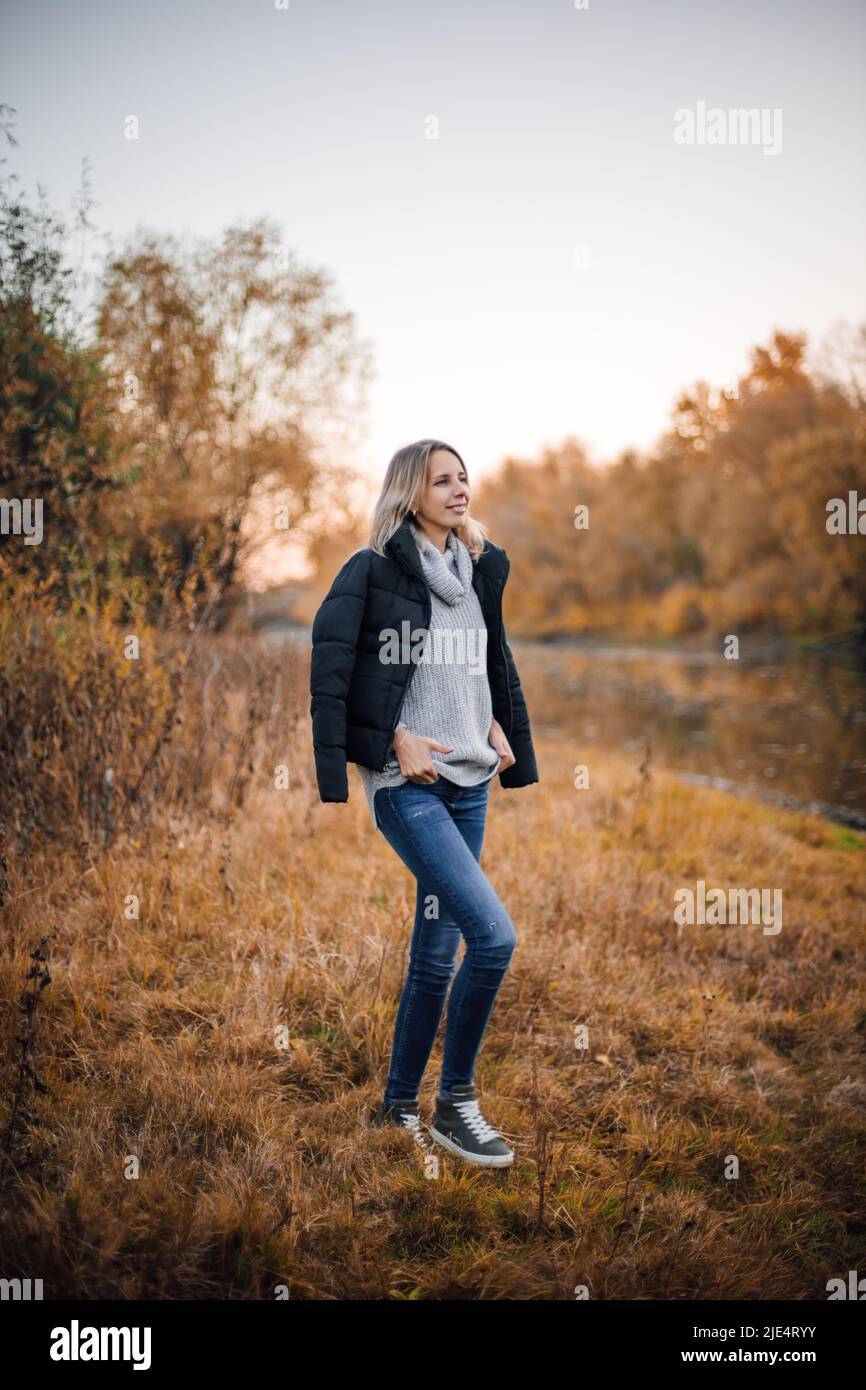 Ritratto a tutta lunghezza di donna in piedi nel prato vicino fiume guardando via sorridente con giacca nera sulle spalle, mani sul maglione con alberi coperti Foto Stock