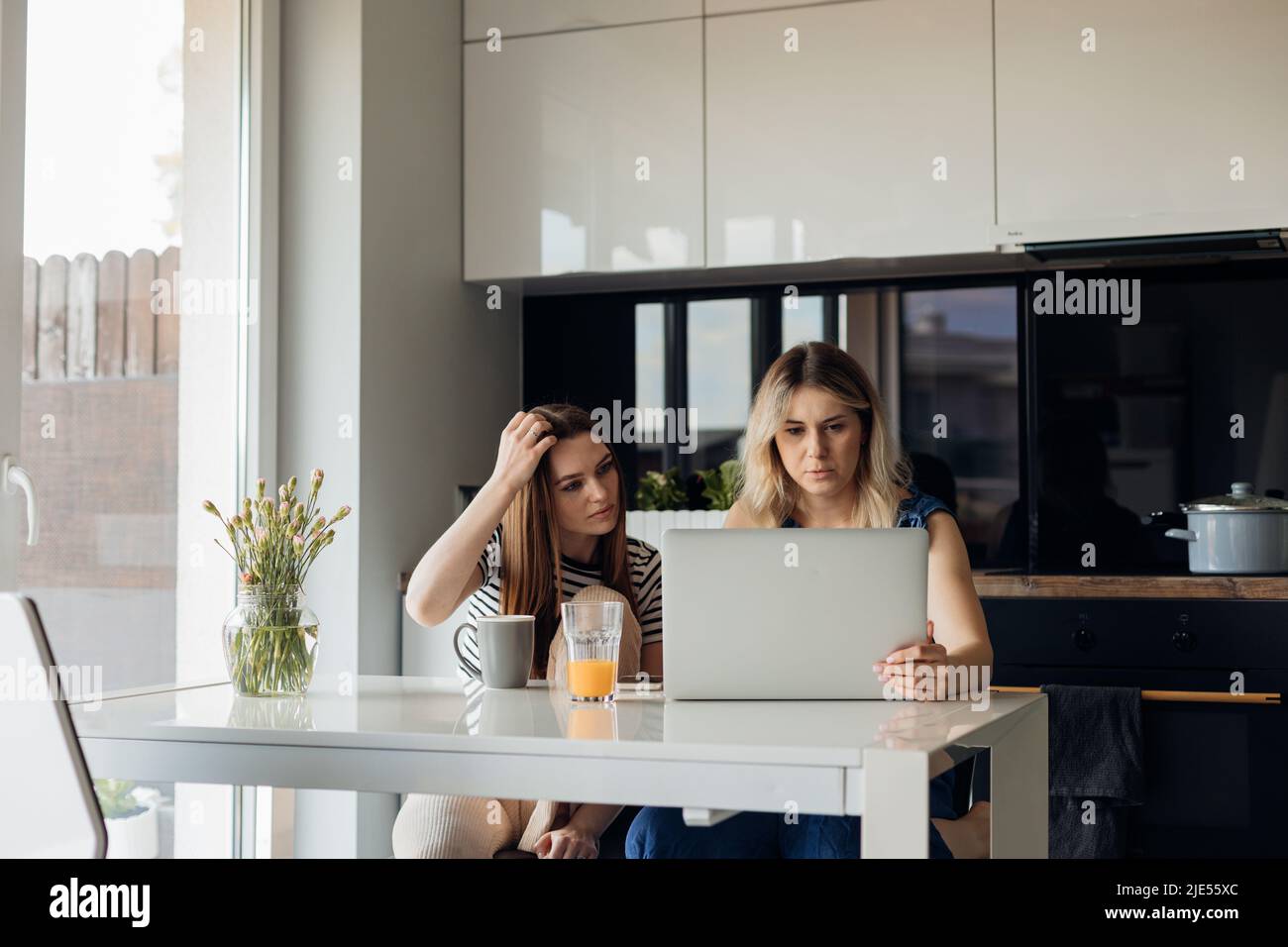 Due donne sedute al tavolo in una cucina moderna e luminosa, che lavorano insieme su un computer portatile e bevono succo di frutta fresco e caffè al mattino. Lavoro indipendente, a distanza Foto Stock