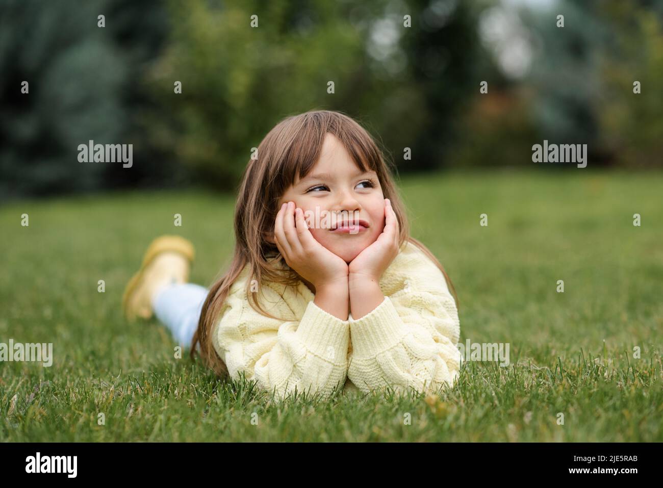 Divertente sorridente bionda bambina di 4-5 anni sdraiata sul prato verde erba all'aperto. Maglione casual a maglia da bambino su sfondo naturale in Foto Stock
