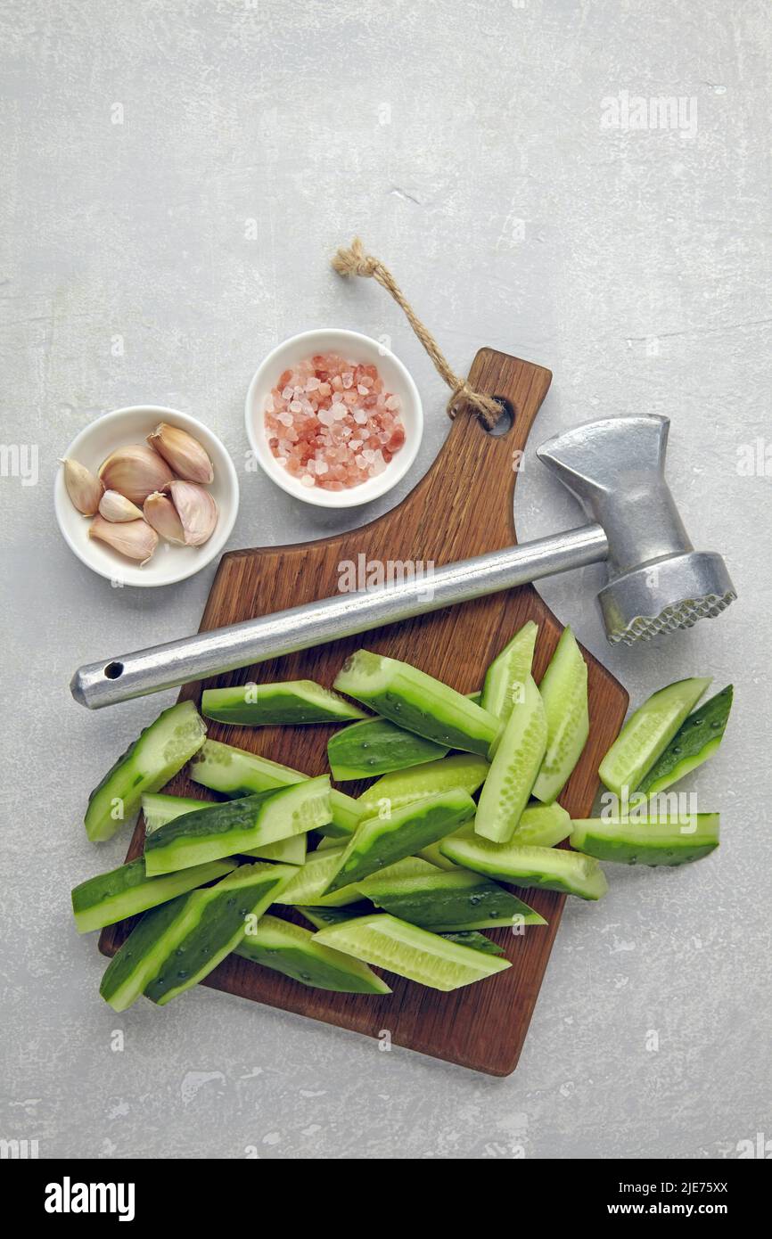 Tagliare cetrioli freschi su un tagliere di legno, martello da cucina, aglio e sale rosa su un tavolo di cemento leggero. Cucina di antipasti da cucum rotto Foto Stock