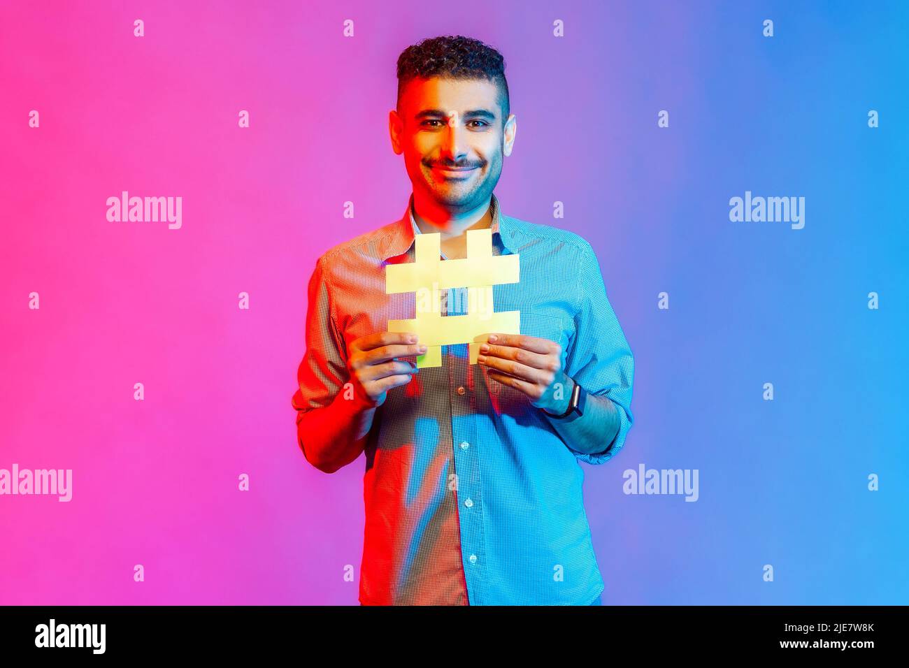 Ritratto di uomo in camicia con il sorriso toothy sul suo volto che tiene hashtag simbolo, consigliando i soggetti popolari, tendenze del Internet. Studio interno girato isolato su colorato sfondo di luce al neon. Foto Stock