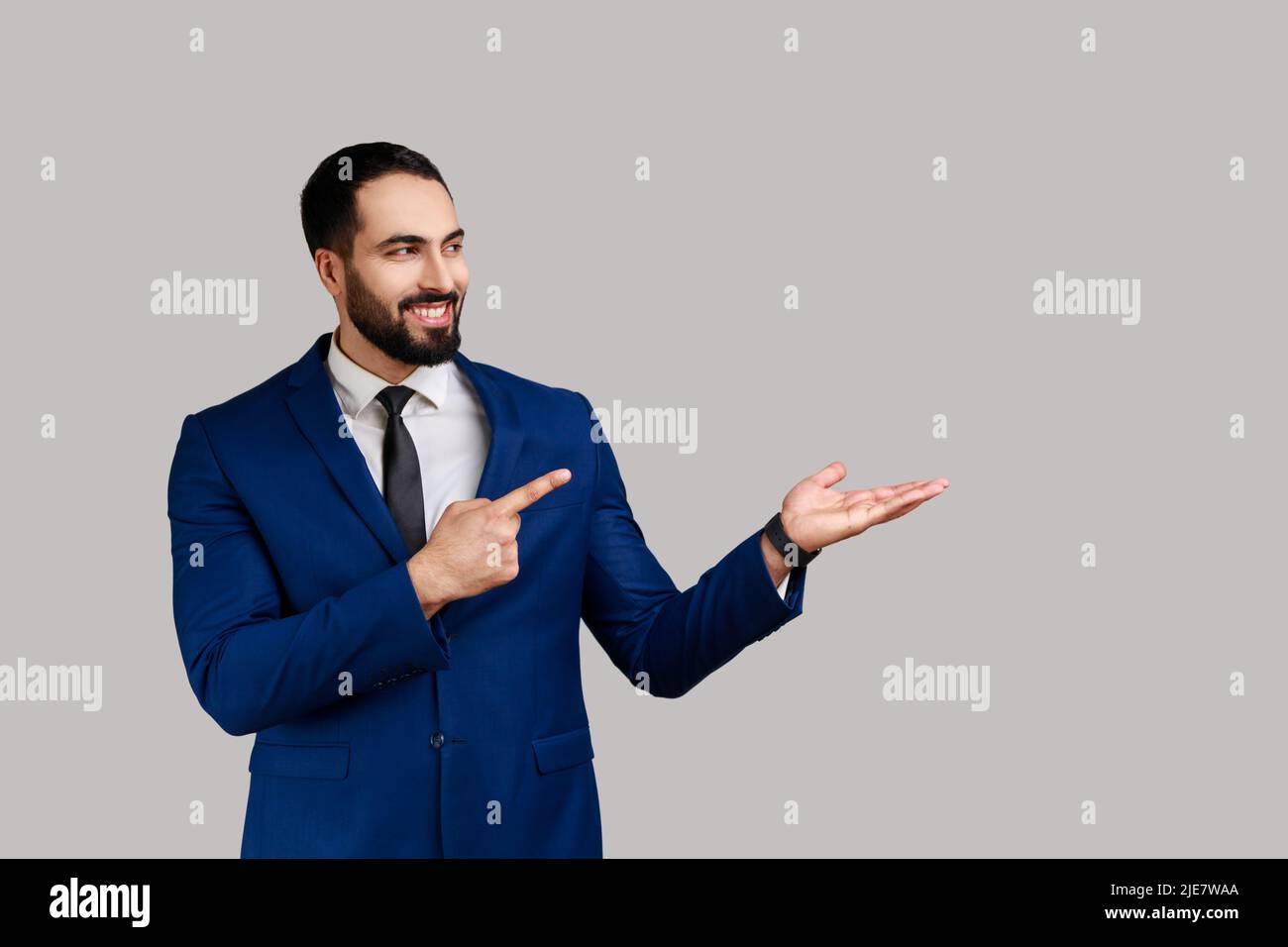 Uomo bearded che presenta lo spazio della copia sul suo palmo e che osserva sorridente, mostrando il posto vuoto per il testo promozionale, indossando il vestito di stile ufficiale. Studio interno girato isolato su sfondo grigio. Foto Stock