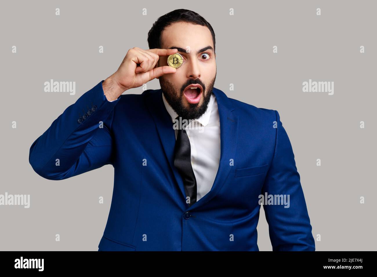 Ritratto di uomo d'affari bearded che copre l'occhio con bitcoin dorato, guardando la macchina fotografica con espressione scioccata, indossando il vestito ufficiale di stile. Studio interno girato isolato su sfondo grigio. Foto Stock