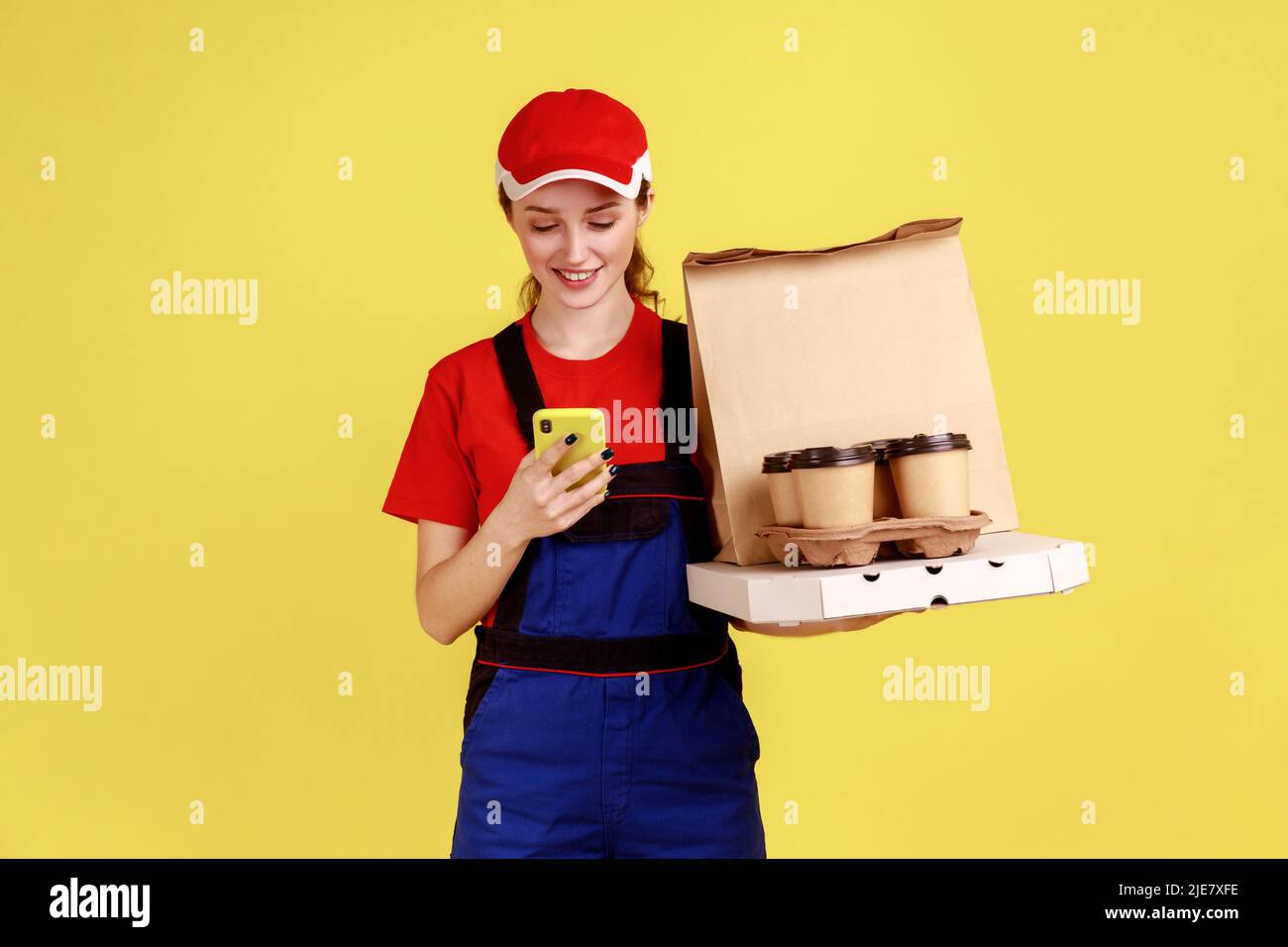 Ritratto di donna con consegna positiva che tiene le scatole di caffè e pizza, utilizzando lo smartphone per controllare l'indirizzo per la consegna, indossare tute e tappo rosso. Studio interno girato isolato su sfondo giallo. Foto Stock