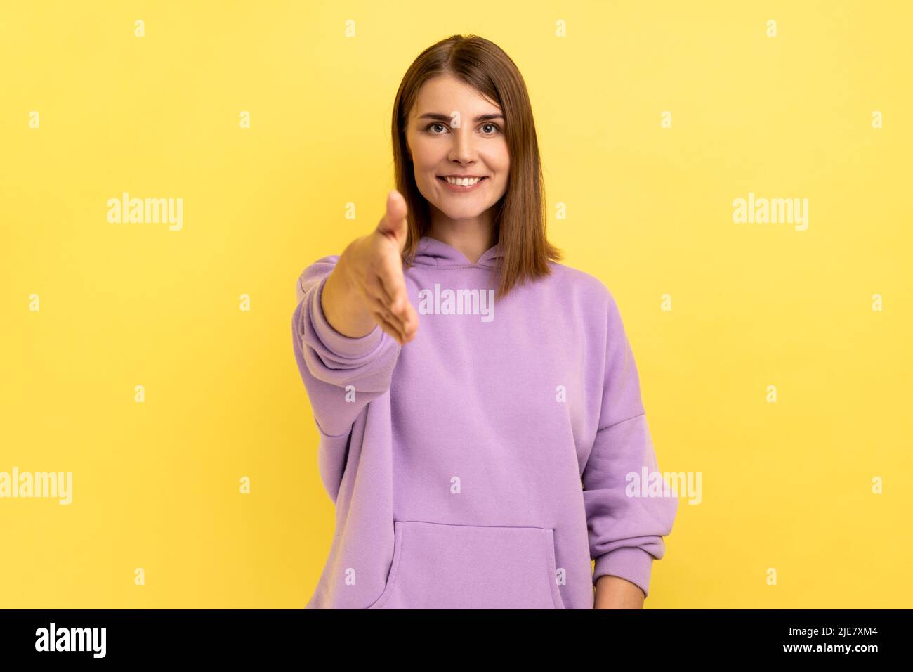 Piacere di conoscerti. Ritratto di felice amichevole donna sorridente che allunga la mano accogliendo gli ospiti, ospitalità, indossando felpa con cappuccio viola. Studio interno girato isolato su sfondo giallo. Foto Stock