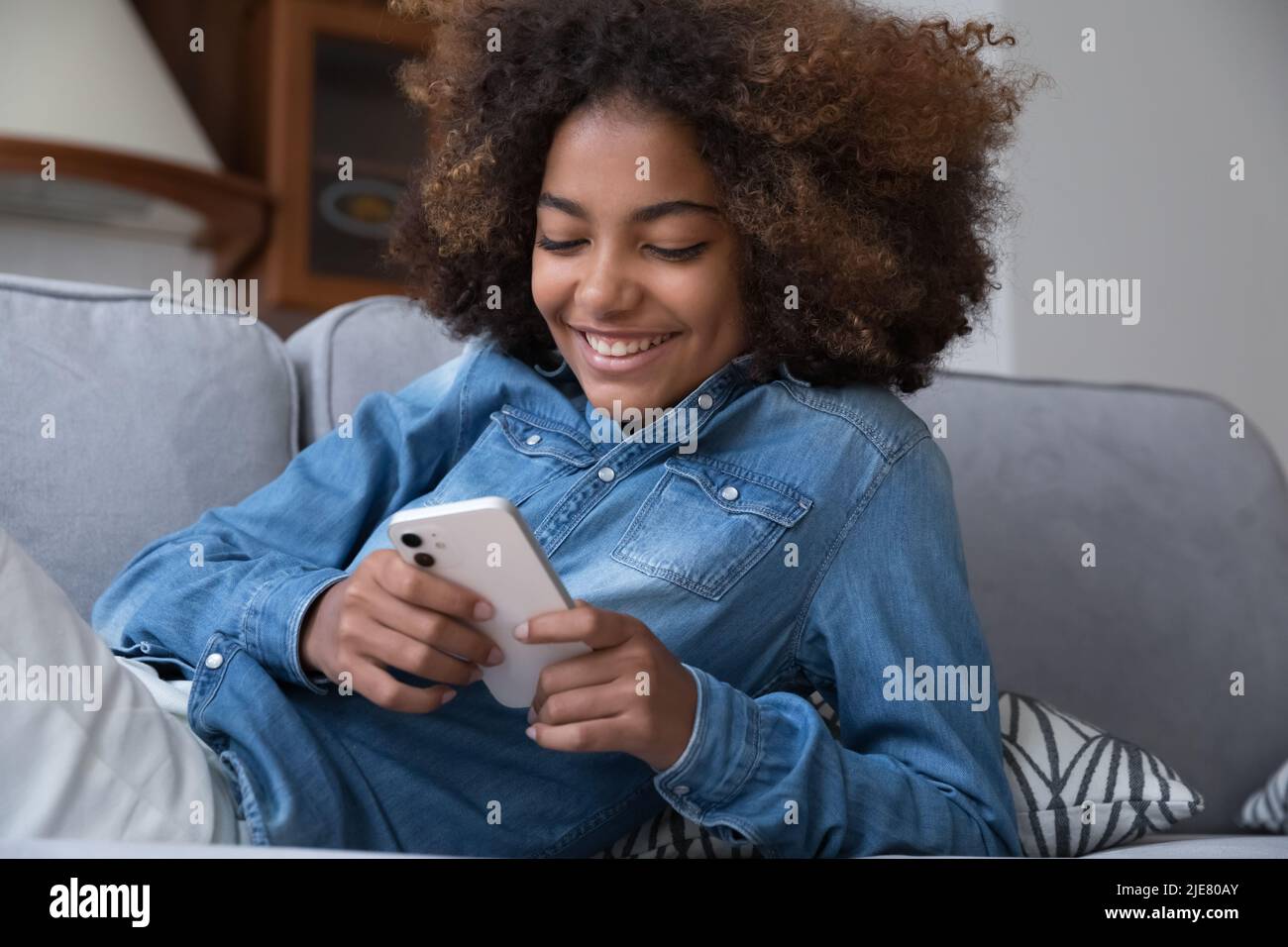 Felice ragazza adolescente africana sedersi sul divano utilizzare smartphone moderno Foto Stock