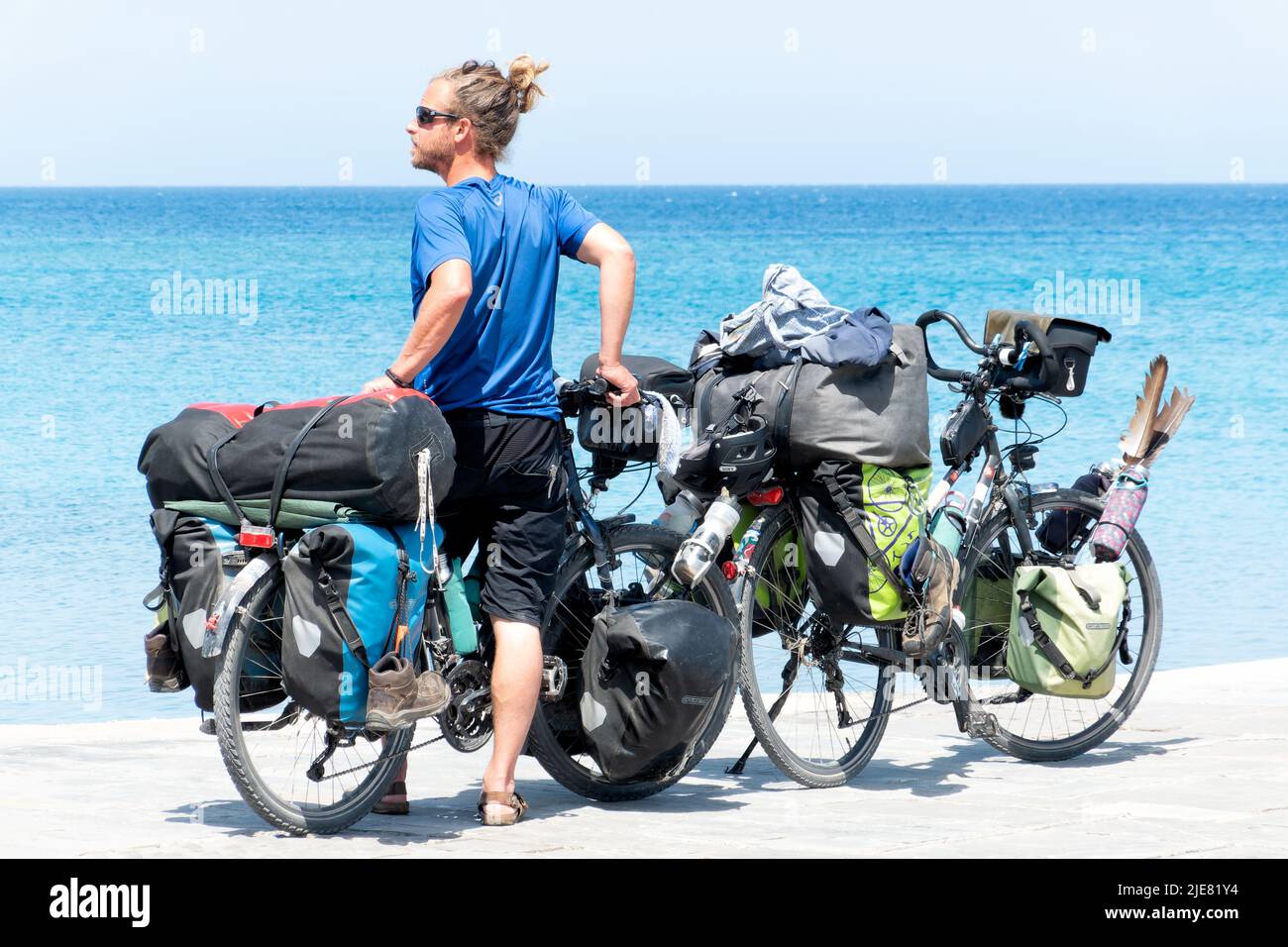 Un uomo si riposa e si alza reggendo due biciclette mentre guarda verso il mare. Le biciclette sono molto cariche di equipaggiamento per una vacanza o un tour in bikepacking Foto Stock