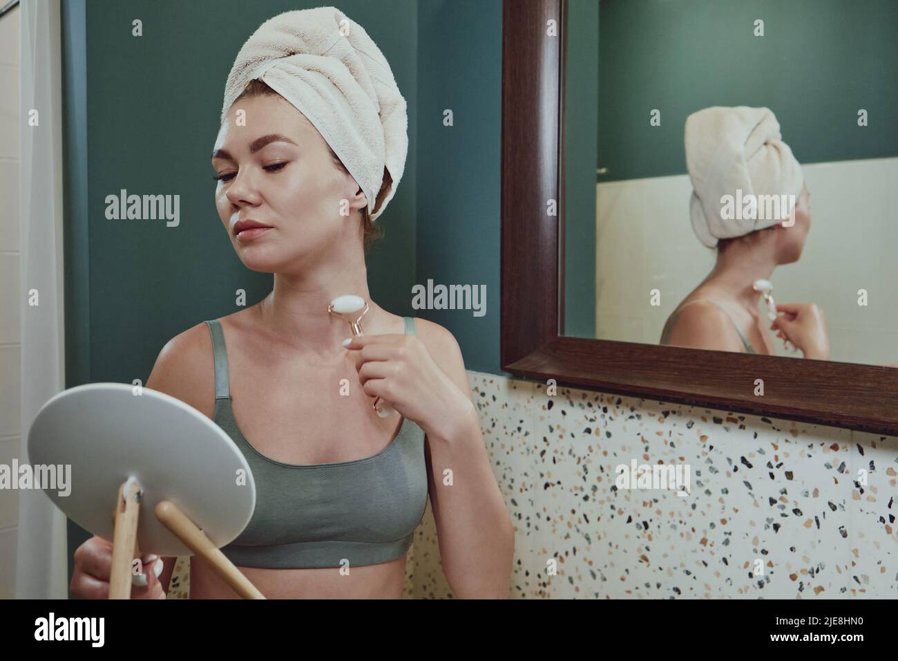 Giovane donna con rullo per il viso in giada per massaggio al collo seduto accanto allo specchio del bagno Foto Stock