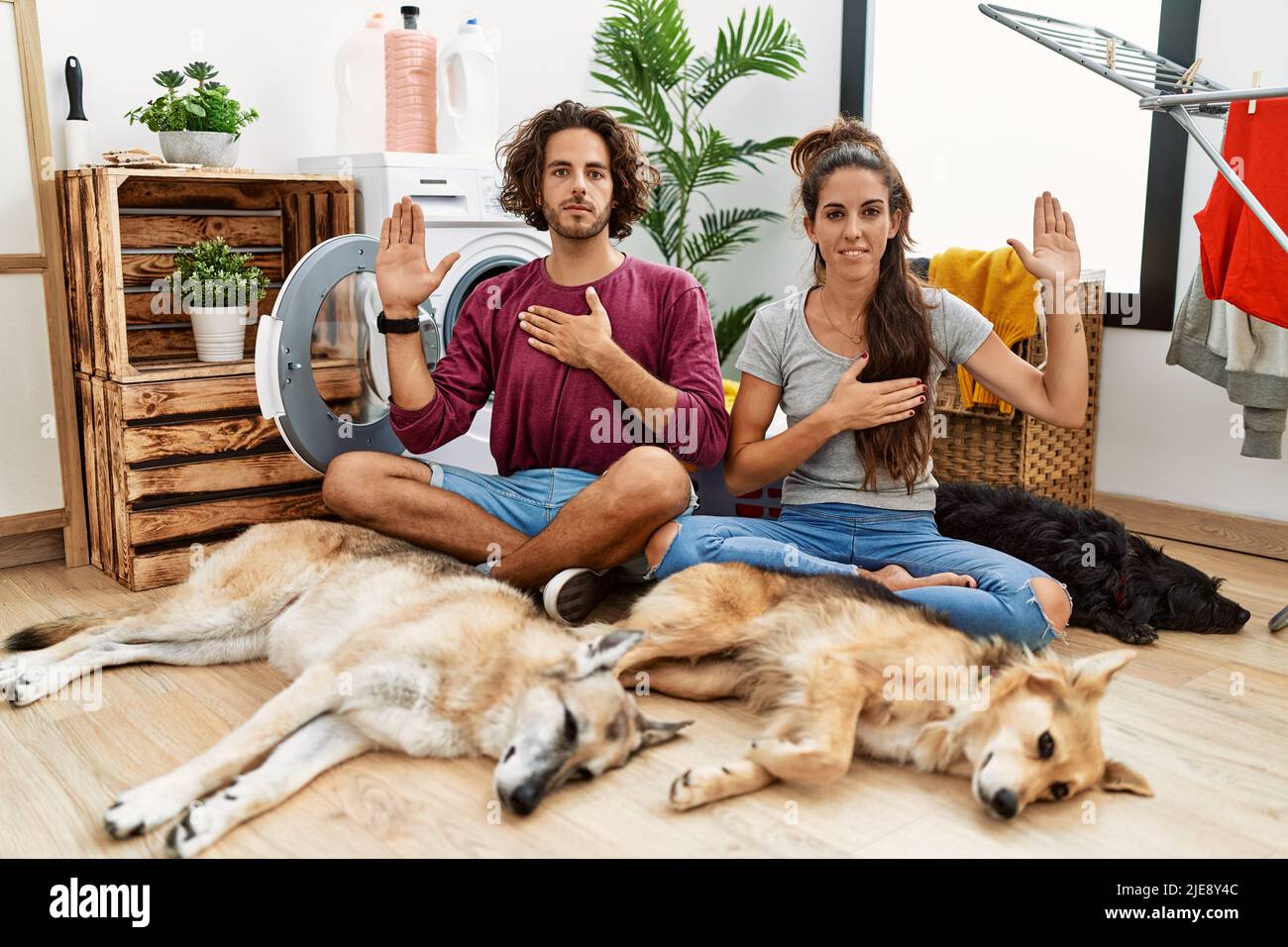 Giovane coppia ispanica che fa lavanderia con cani giurando con mano sul petto e palma aperta, facendo una promessa di lealtà giuramento Foto Stock