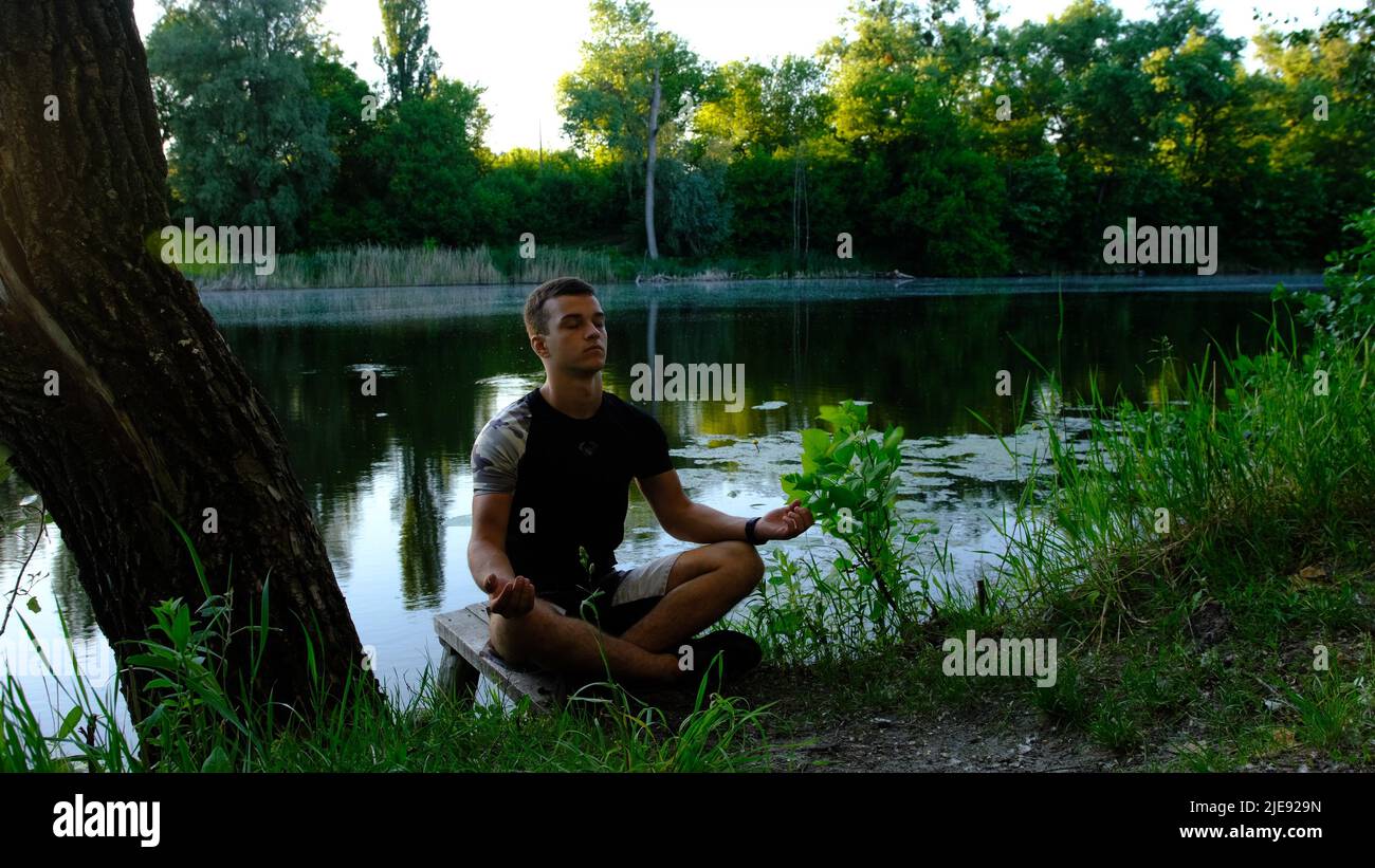 Sportivo bel giovane caucasico che lavora sul lago nel parco, seduto in squat, facendo metà loto, Padangustha Padma Utkatasana, mani in Namaste, f Foto Stock